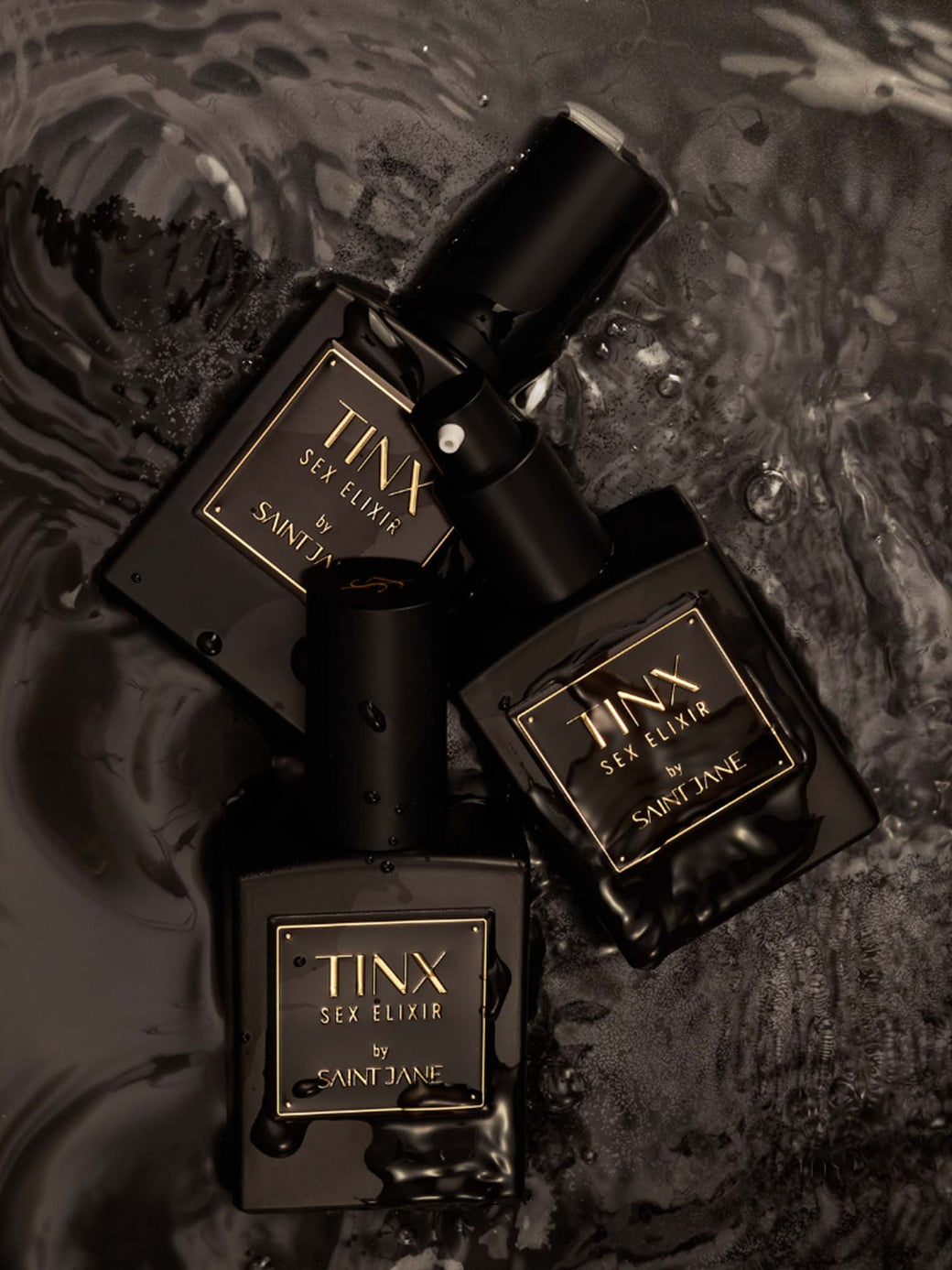 Three black TINX Sex Elixir by Saint Jane bottles on a dark textured surface.