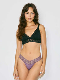 A woman modeling a two-piece lingerie set consisting of a black lace bra and matching purple lace thong, set against a plain light gray background.
