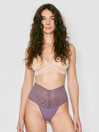 A woman modeling a two-piece lingerie set against a plain light gray studio background. The set includes a beige lace-trim bralette and high-waisted purple lace briefs.