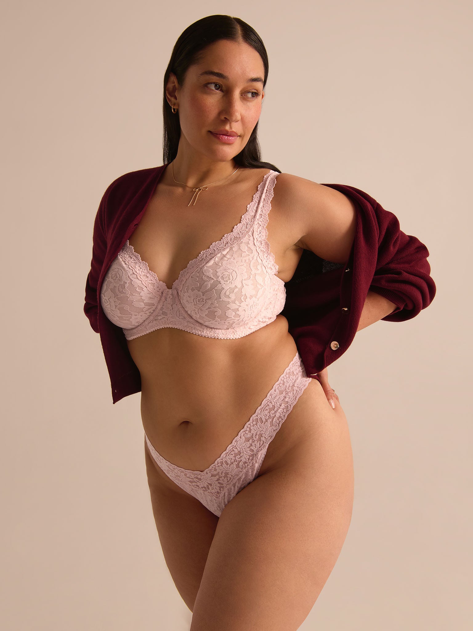 Woman wearing a white lace bra and panties set with a burgundy jacket over her shoulders on a beige background.